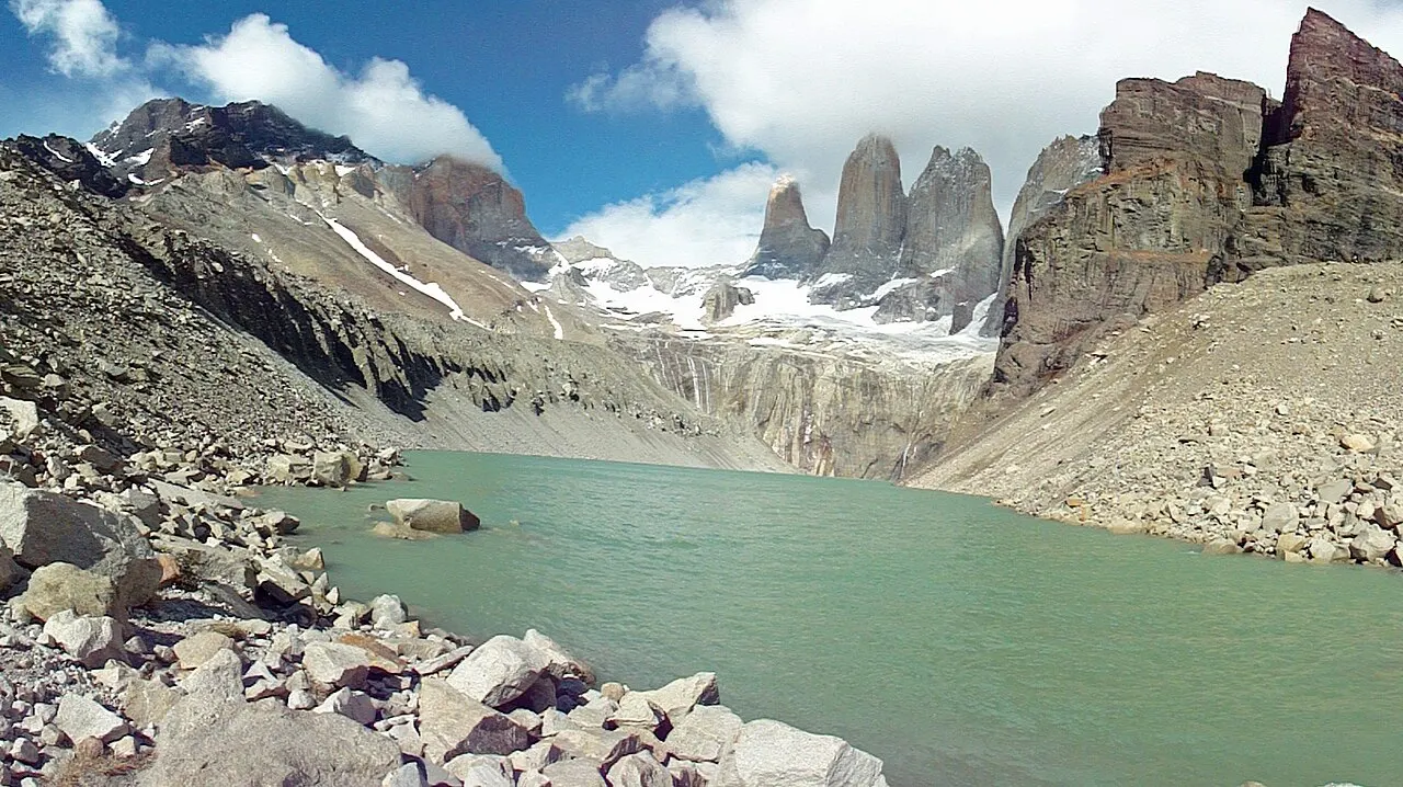 As três torres de granito do Paine no fundo do vale, acima de uma lagoa verde cercada de pedras soltas, sob um céu com nuvens.