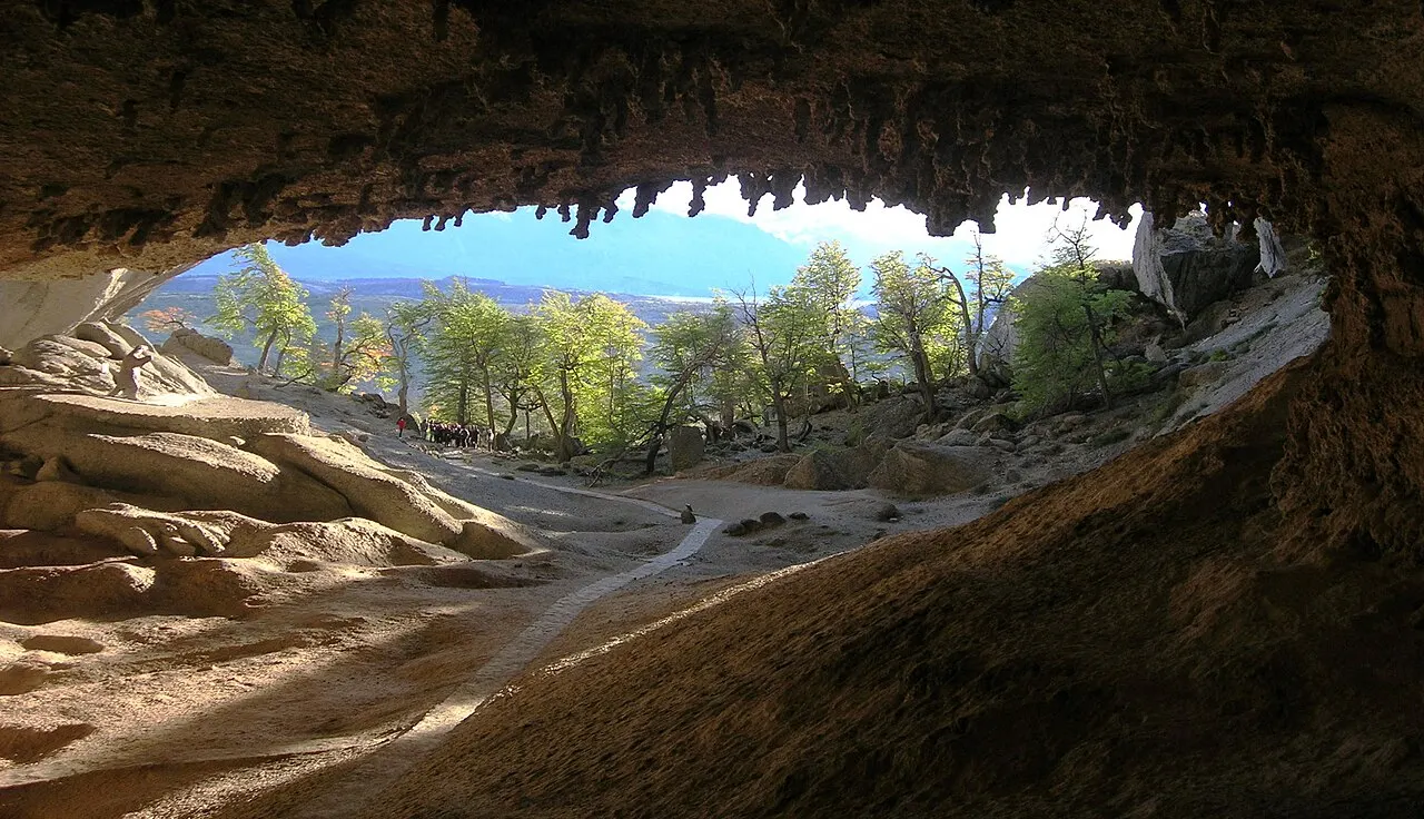 O interior da Cueva del Milodón olhando para a saída: o teto de rocha irregular enquadra árvores e um céu claro do lado de fora.