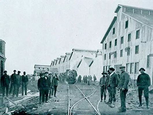 Fotografia em preto e branco de cerca de 1920 do frigorífico Bories: galpões de madeira em fila e um grupo de homens de chapéu sobre os trilhos.