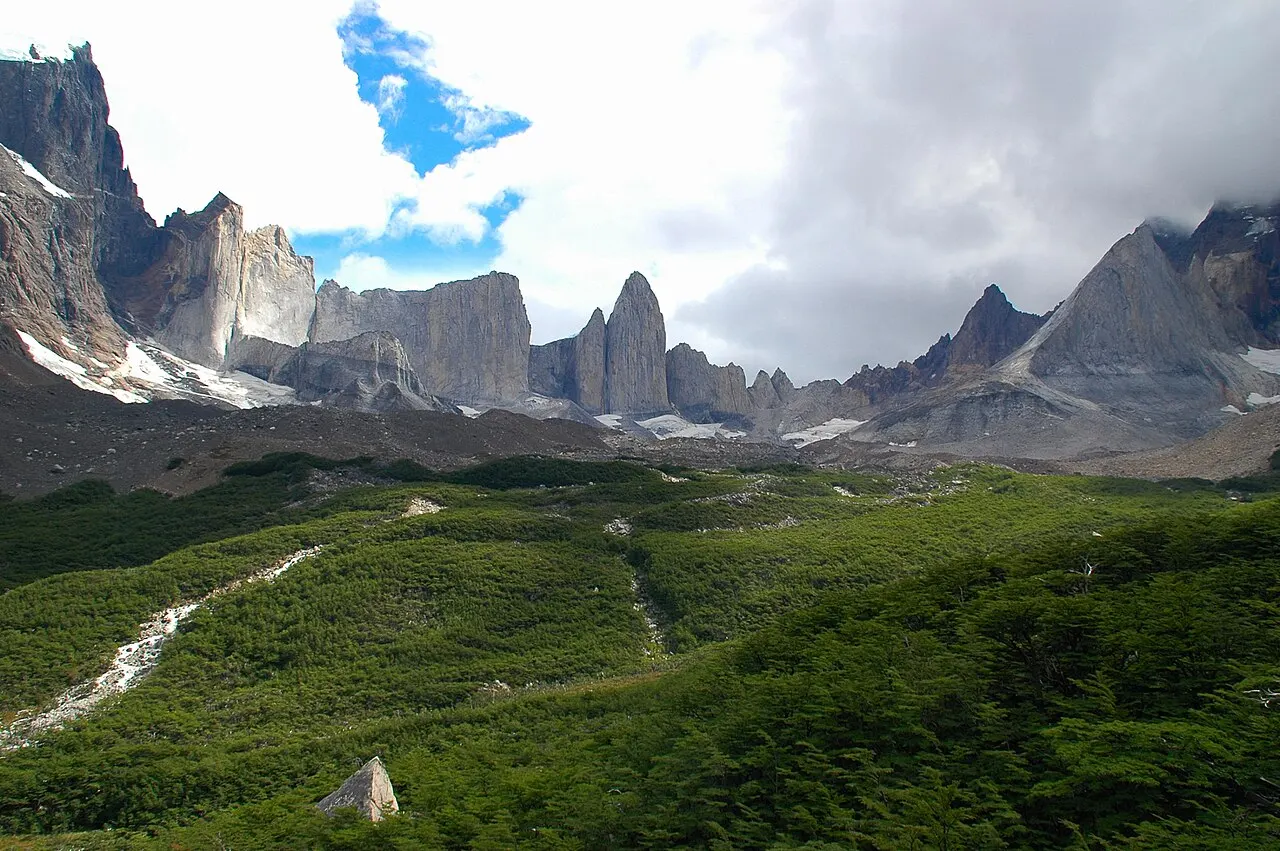O Valle del Francés coberto de mato verde, com uma trilha descendo a encosta e a muralha de picos de granito do Paine ao fundo.