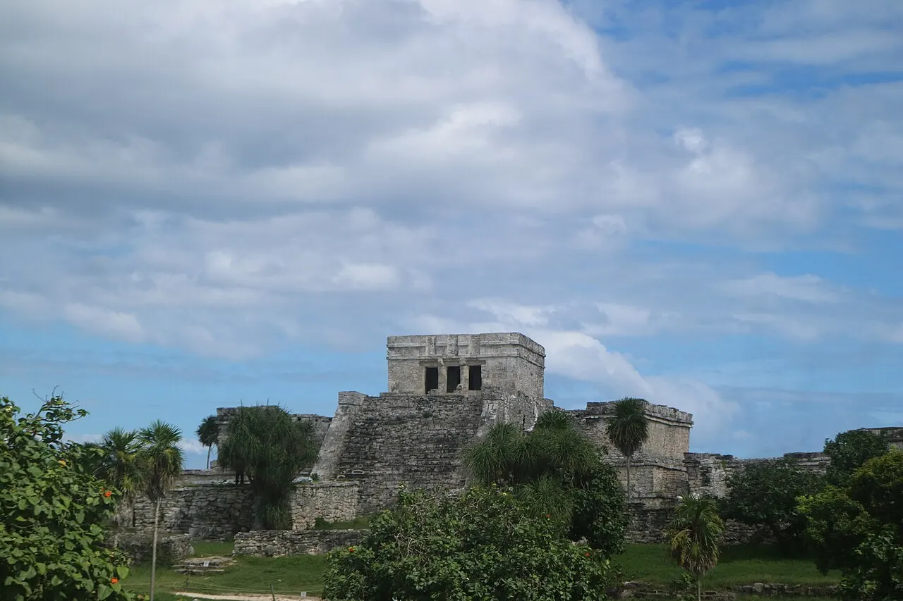 El Castillo, a estrutura de pedra do sítio de Tulum, vista de longe entre a vegetação, sob céu com nuvens.