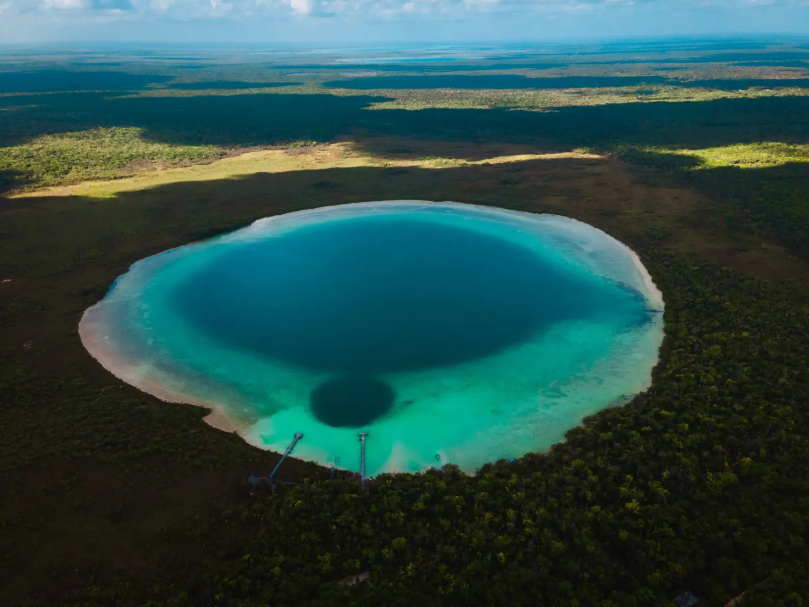 Lagoa circular de água turquesa com uma área mais escura no centro, dois píeres e uma floresta ao redor.