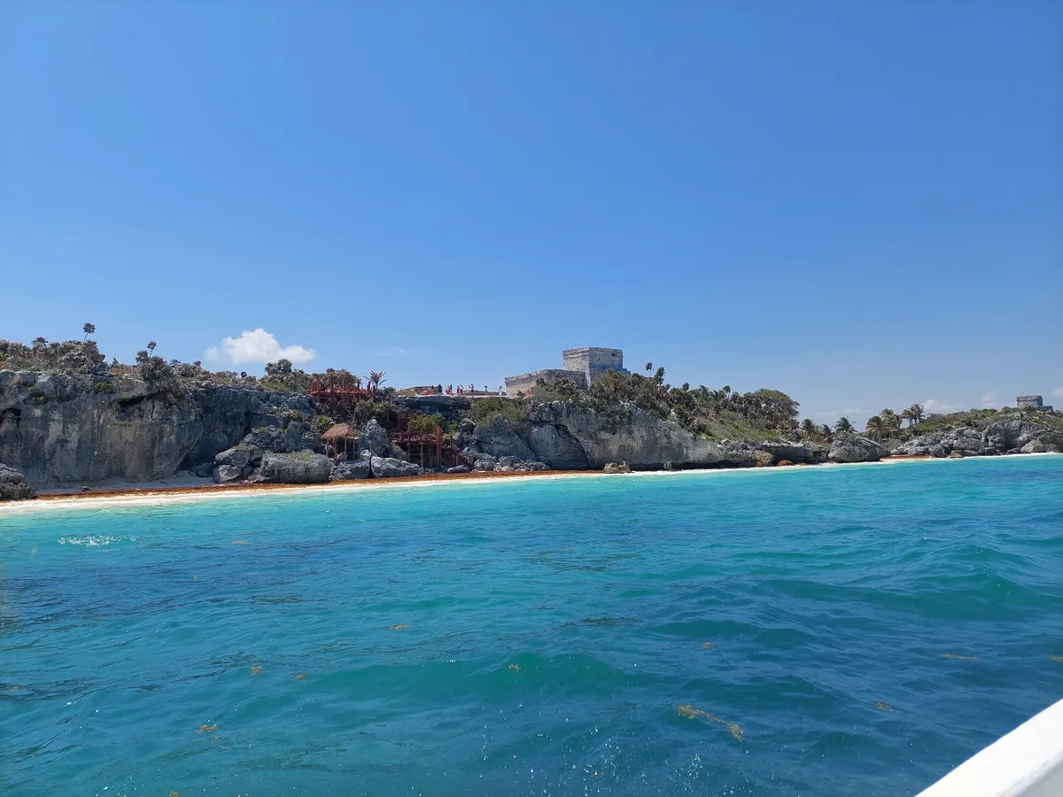 Mar azul diante de falésias, vegetação e ruínas de Tulum; faixa da praia e pequenos visitantes ao longe, com parte do barco no canto.