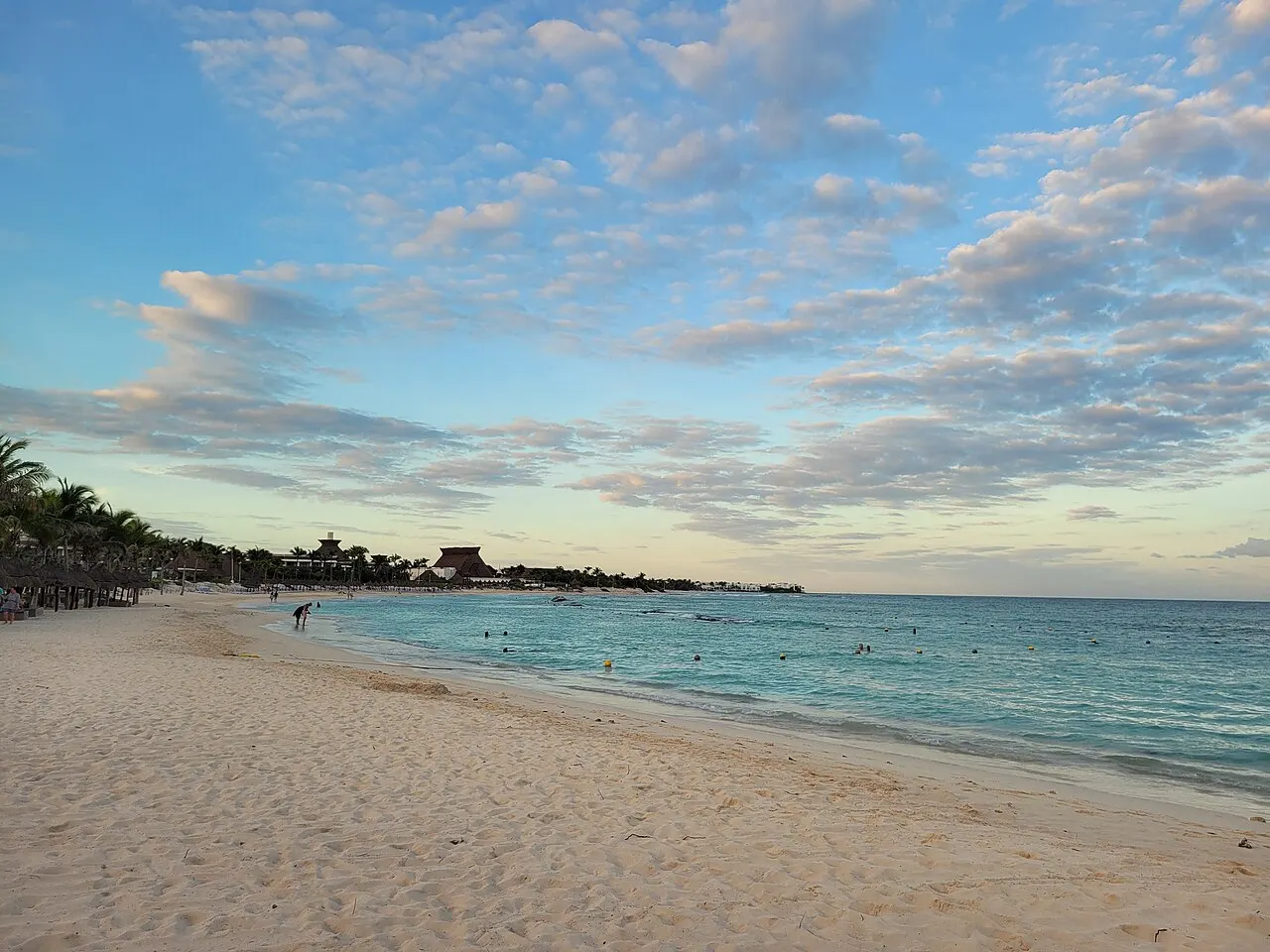 Praia de areia clara em Tulum com mar de água turquesa, palapas de palha ao longe e céu com nuvens espalhadas ao entardecer.