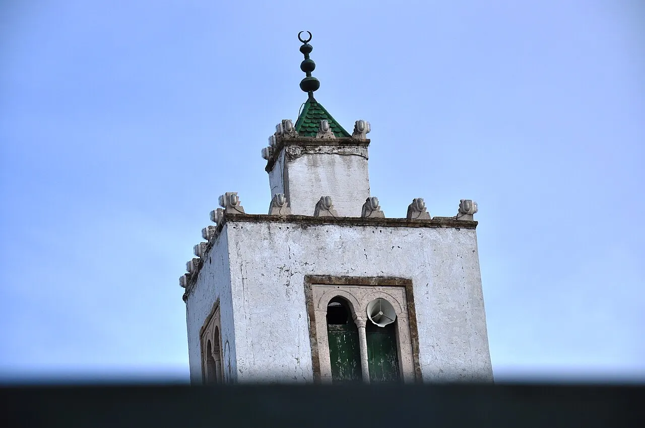 El minarete blanco de la mezquita Sidi Mahrez, en Túnez, con ventanas en arco y tejado verde, visto desde abajo.