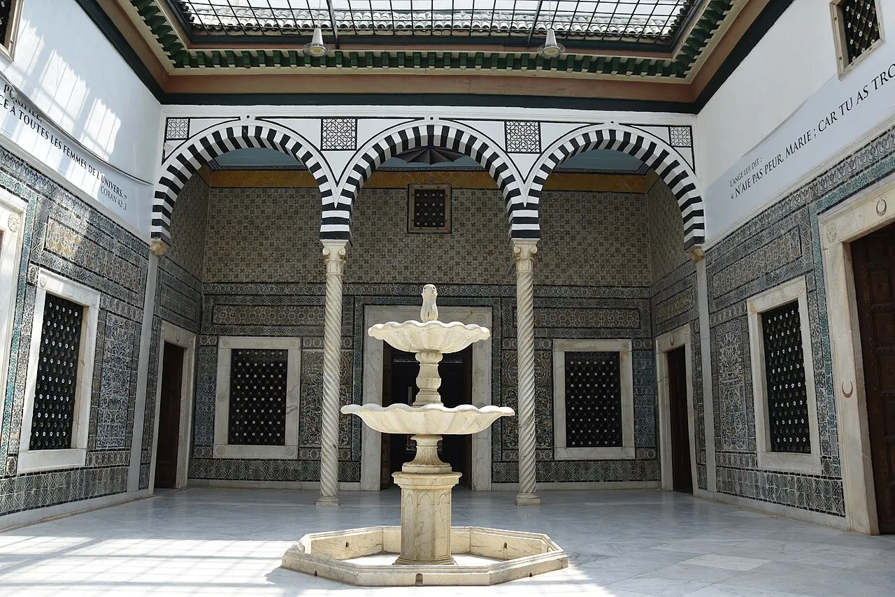Pátio interno do Museu do Bardo com arcos em ferradura listrados de preto e branco, paredes cobertas de azulejos e uma fonte de mármore ao centro.