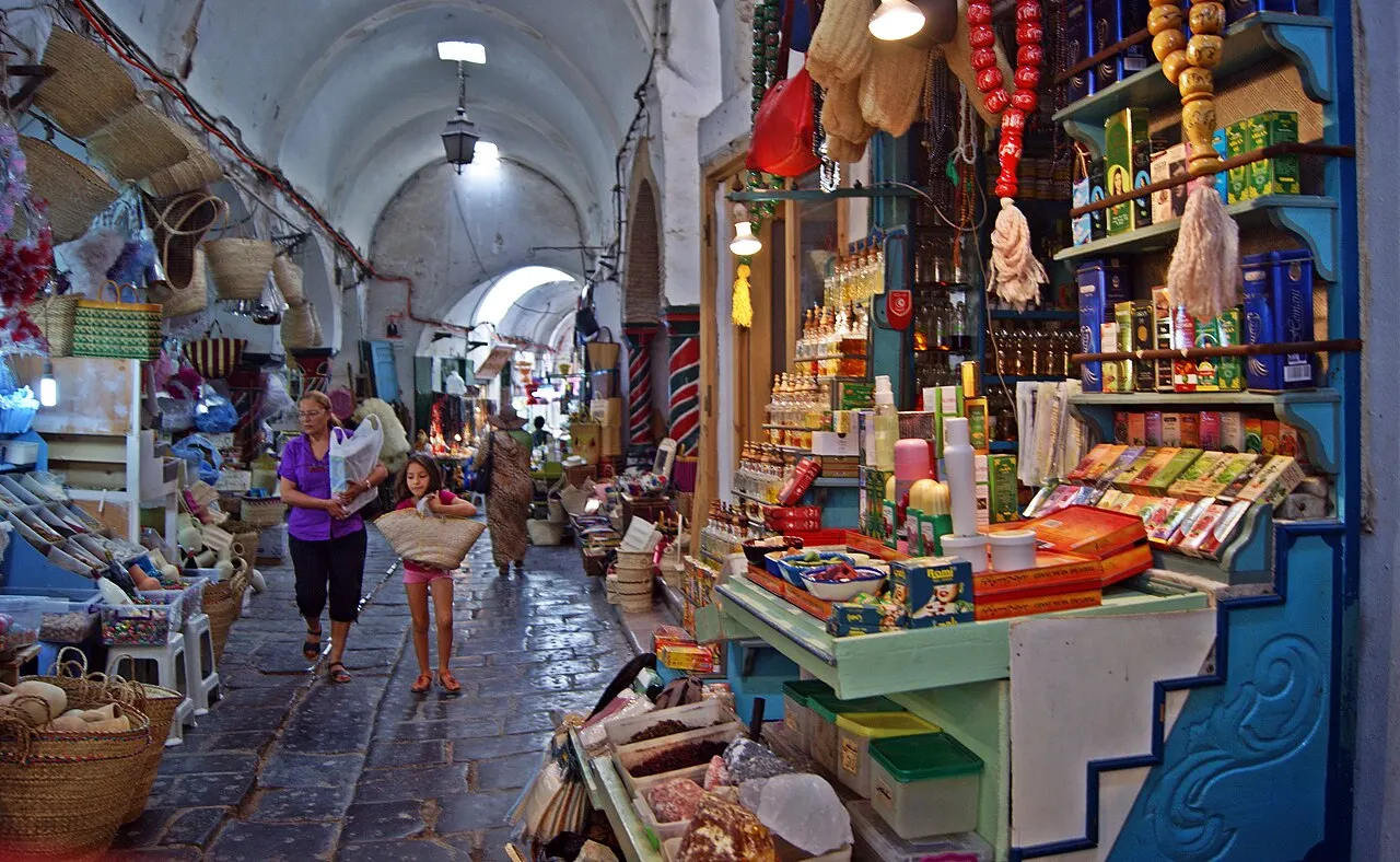 Corredor coberto do souk da medina de Túnis, com bancas de mercadorias coloridas e pessoas caminhando.