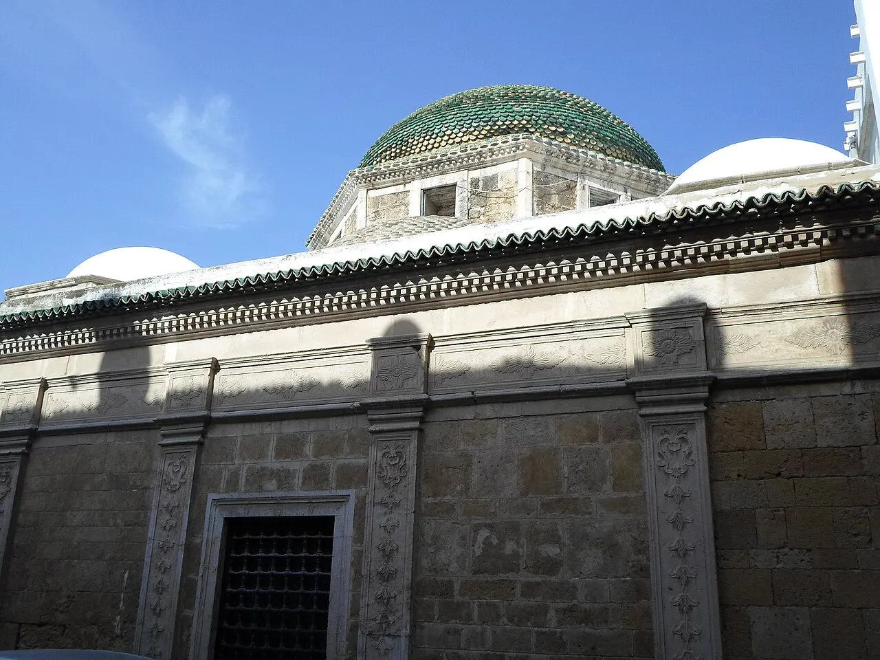Fachada lateral do Tourbet el Bey, com friso decorado e cúpula verde e branca acima, sob céu azul.