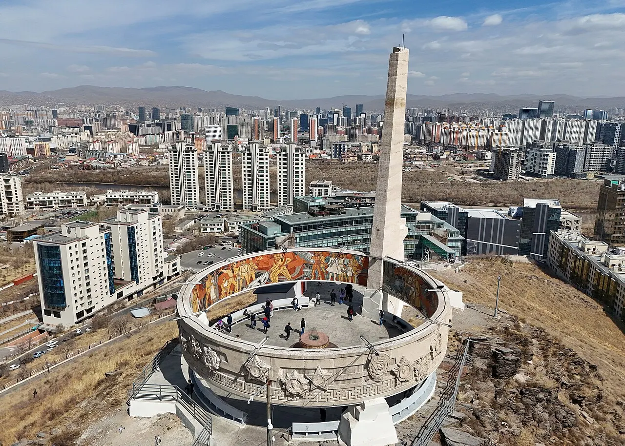 Vista aérea do Memorial Zaisan, com um obelisco alto no centro de uma plataforma circular decorada com um mural colorido, e Ulan Bator ao fundo.