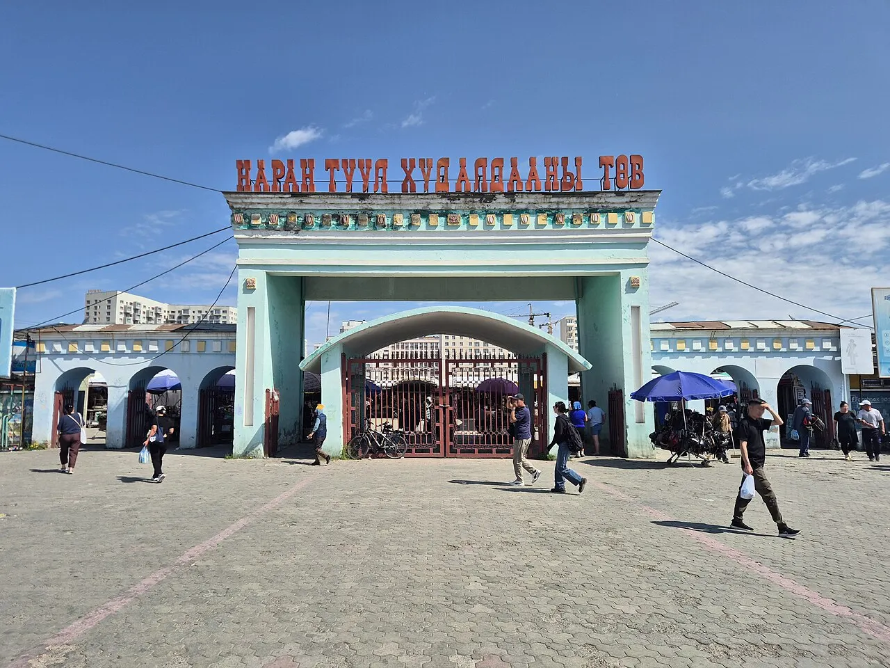 Portão de entrada do Mercado Narantuul, em Ulan Bator, com letreiro vermelho e arco, e pessoas passando.