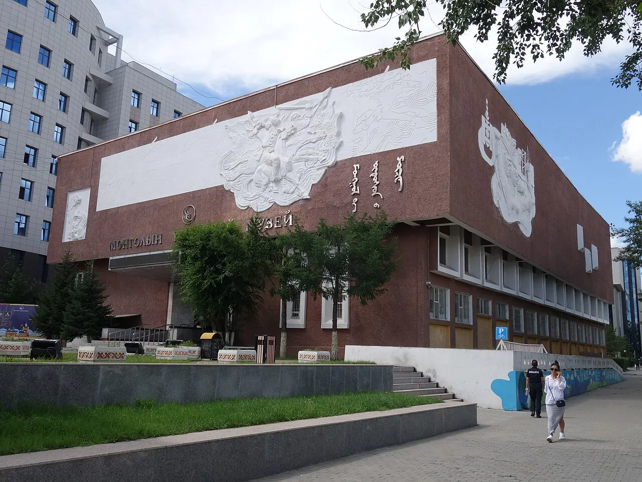 Fachada marrom do Museu Nacional da Mongólia, com grandes relevos brancos de cavaleiros e escrita mongol tradicional, árvores na entrada.