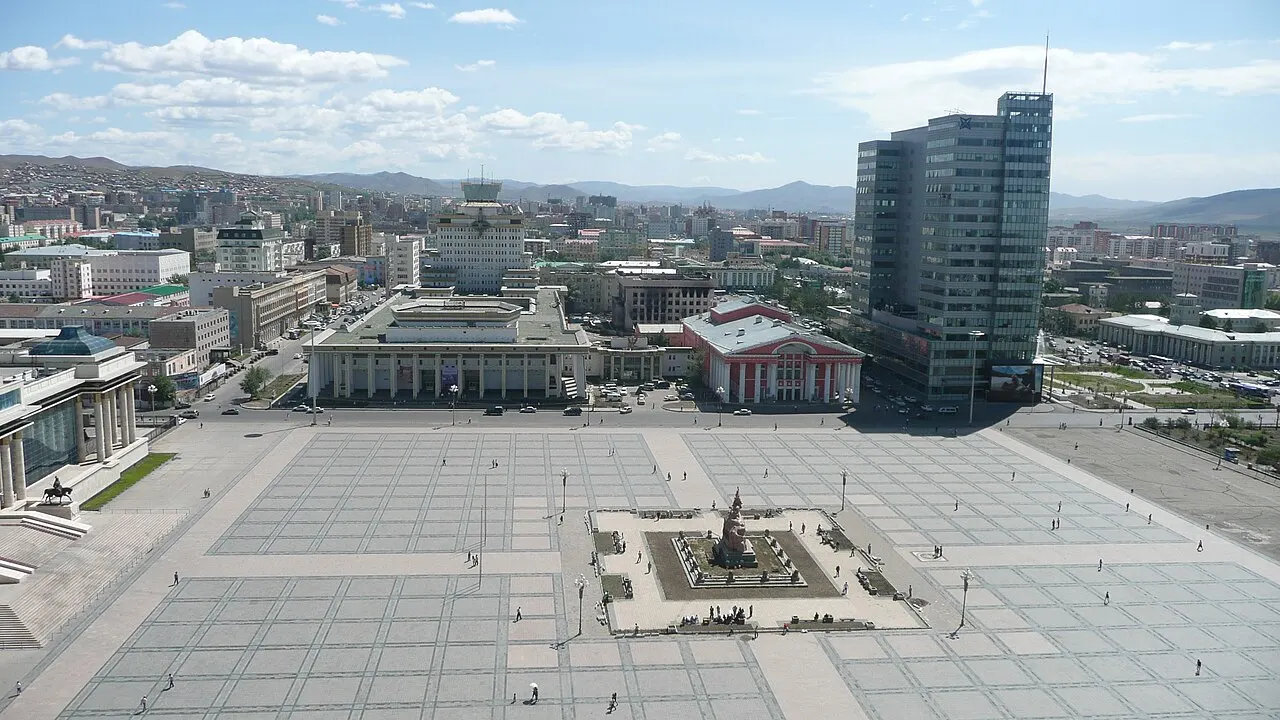 Vista elevada da Praça Sükhbaatar, com um monumento central, prédios governamentais com colunas e uma torre de vidro moderna ao fundo.