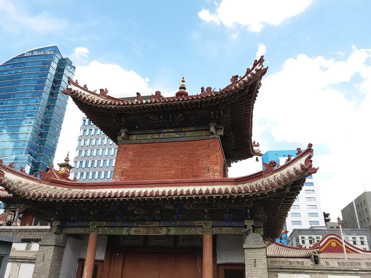 Pavilhão de telhados curvos do Templo-museu Choijin Lama, com torres de vidro de Ulan Bator ao fundo.