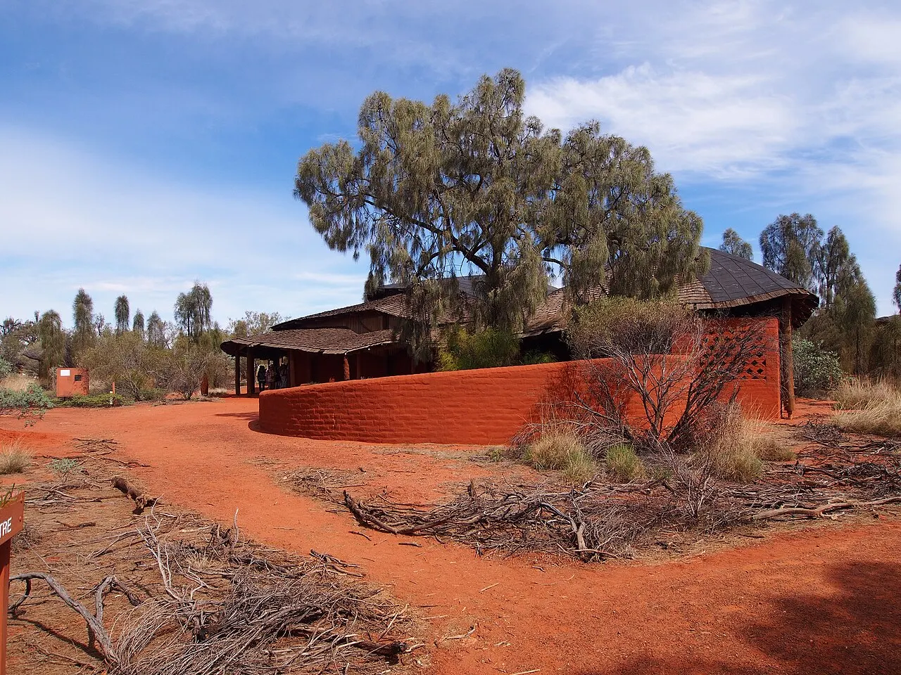 Edifício baixo de terra vermelha do centro cultural de Uluru-Kata Tjuta, com uma árvore alta e pessoas pequenas sob a varanda.