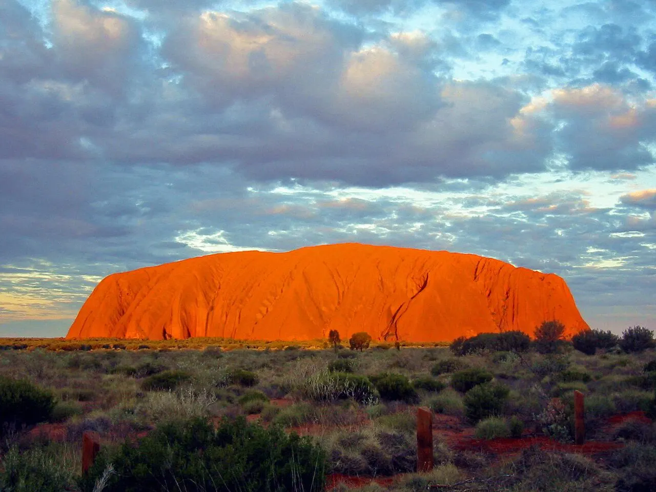 Monólito de Uluru avermelhado pelo pôr do sol, sobre a planície de arbustos, sob céu com nuvens.
