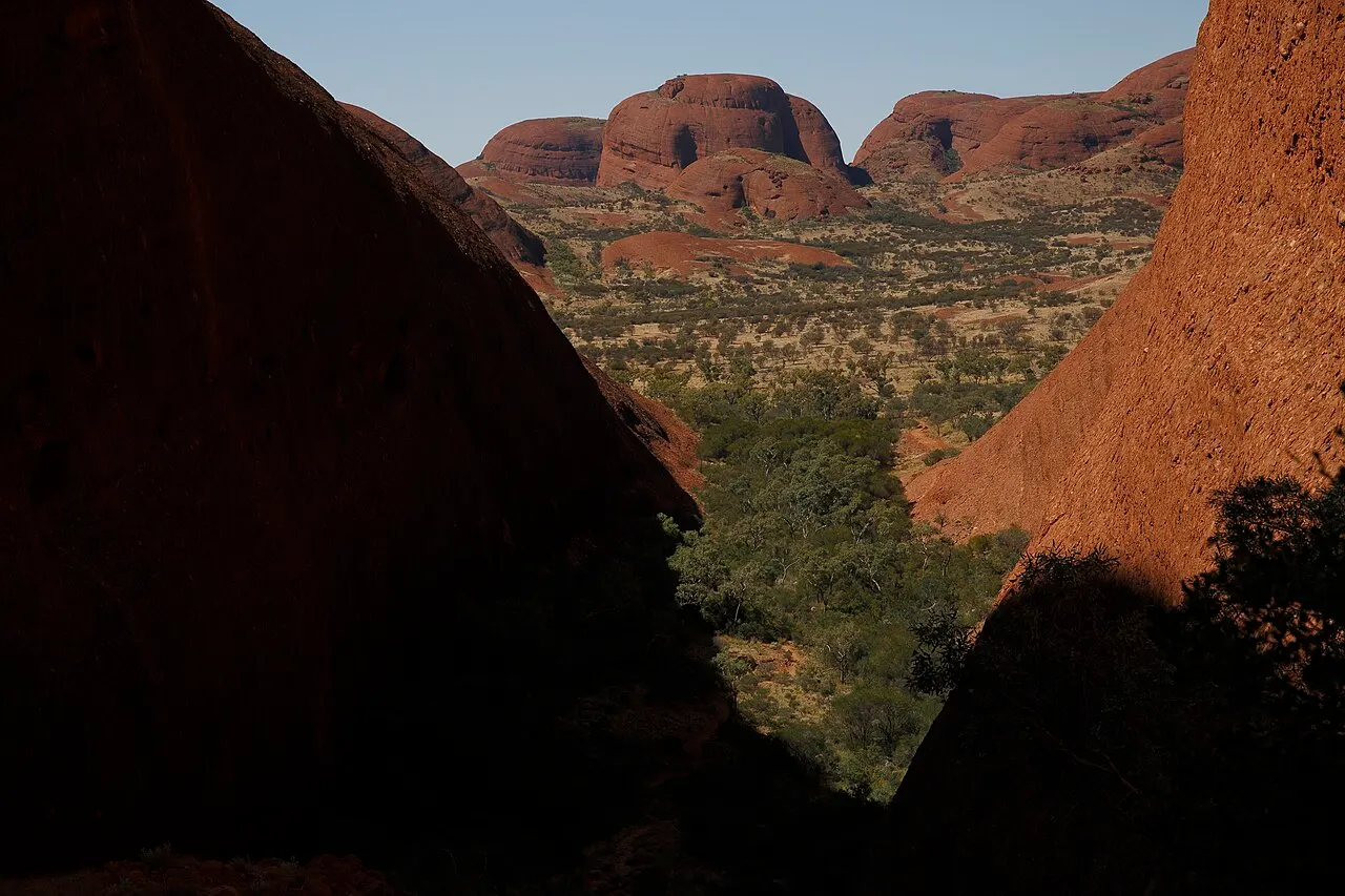 Vista do mirante de Karingana entre duas paredes de rocha vermelha de Kata Tjuta, com o vale arborizado embaixo e mais cúpulas arredondadas ao fundo.