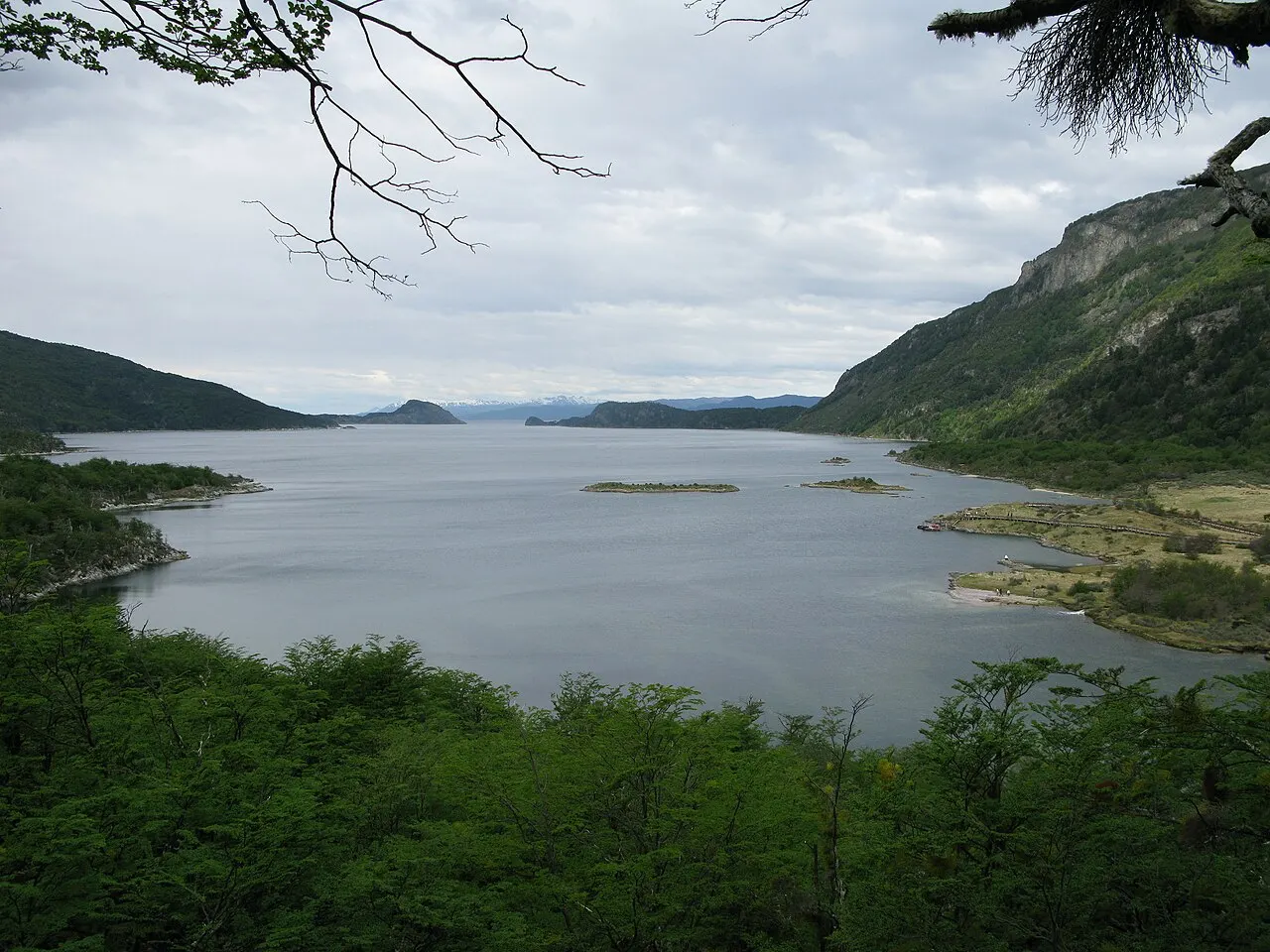A Bahía Lapataia, com água calma, ilhotas e morros cobertos de mata ao fundo, sob céu nublado.