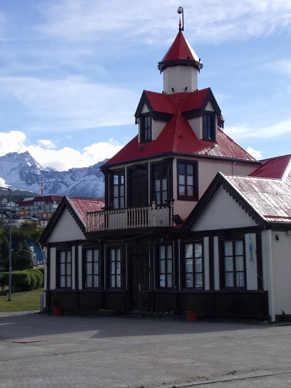 A Casa Beban, de madeira escura, com telhado vermelho e uma torre pequena, e uma montanha ao fundo.