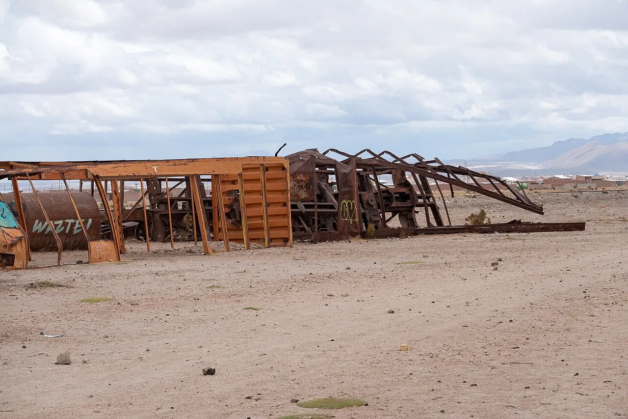 Vagões e estruturas de ferro enferrujadas e desmontadas no cemitério de trens de Uyuni, sobre chão de areia clara, com montanhas e céu nublado ao fundo.