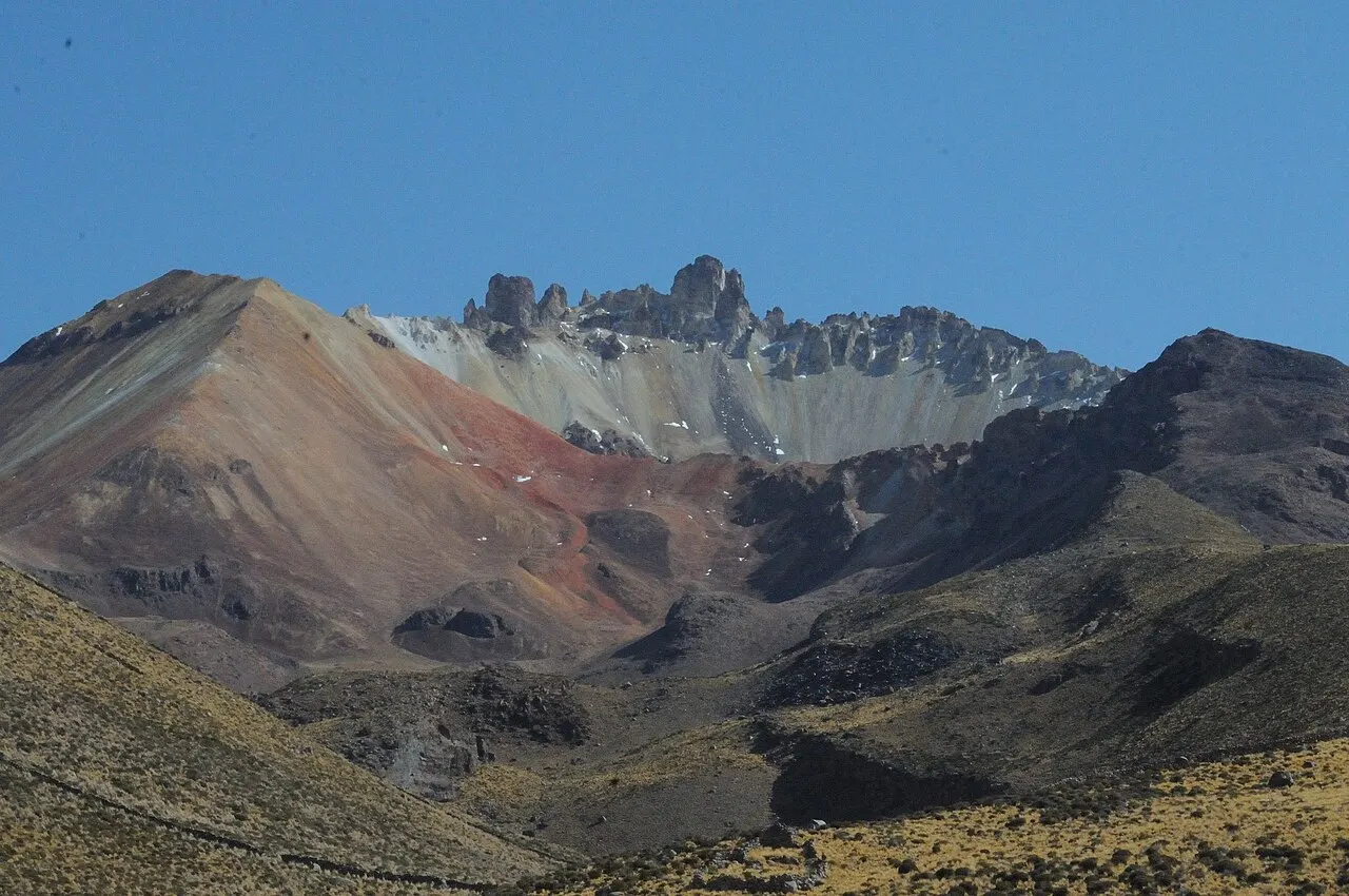 As encostas do vulcão Tunupa em faixas de ocre, vermelho, cinza e creme, com pináculos escuros na borda da cratera e manchas de neve, sobre o altiplano de capim amarelo em primeiro plano.