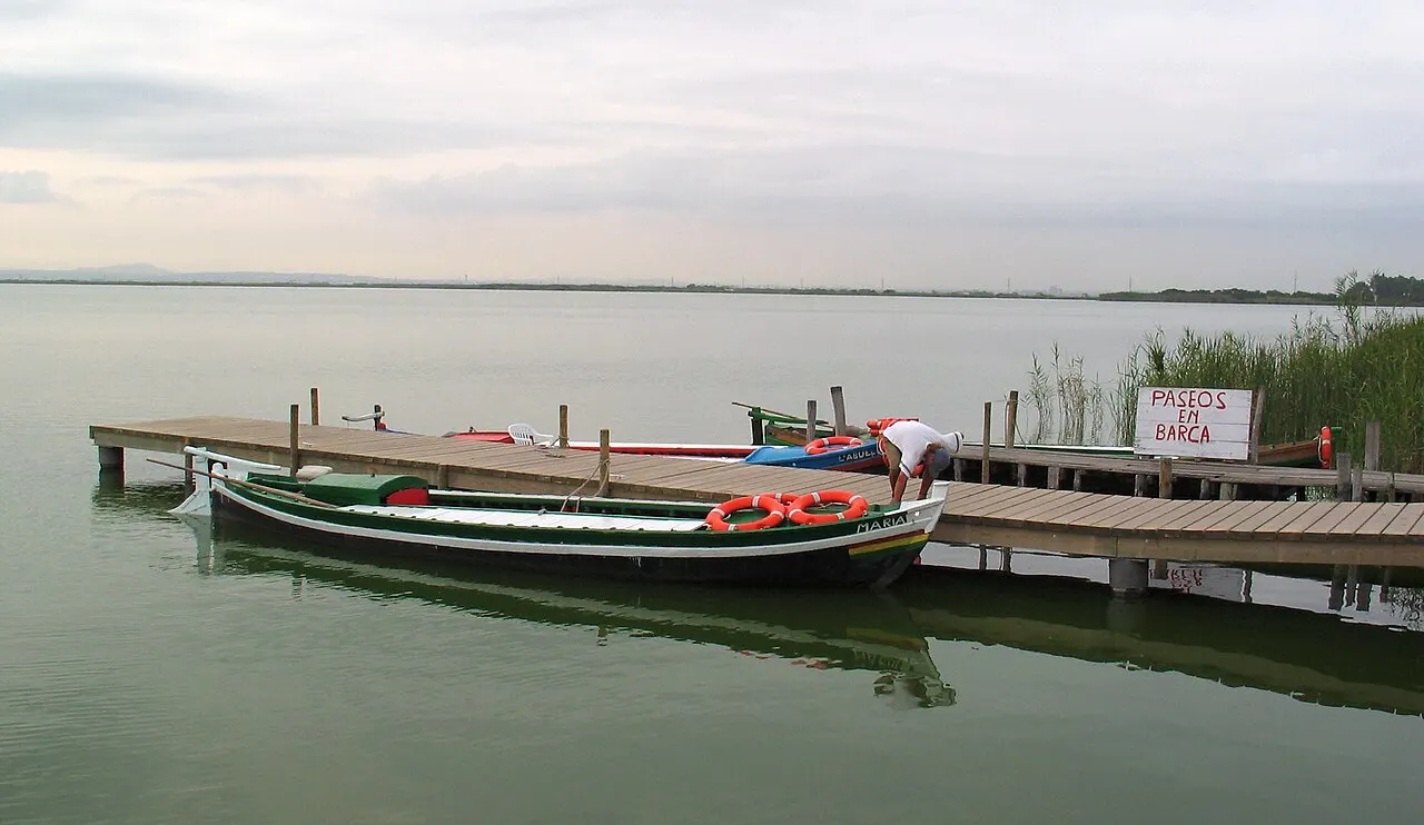 Barcas tradicionais de madeira amarradas a um embarcadouro na lagoa calma da Albufera, com juncos e um céu claro.