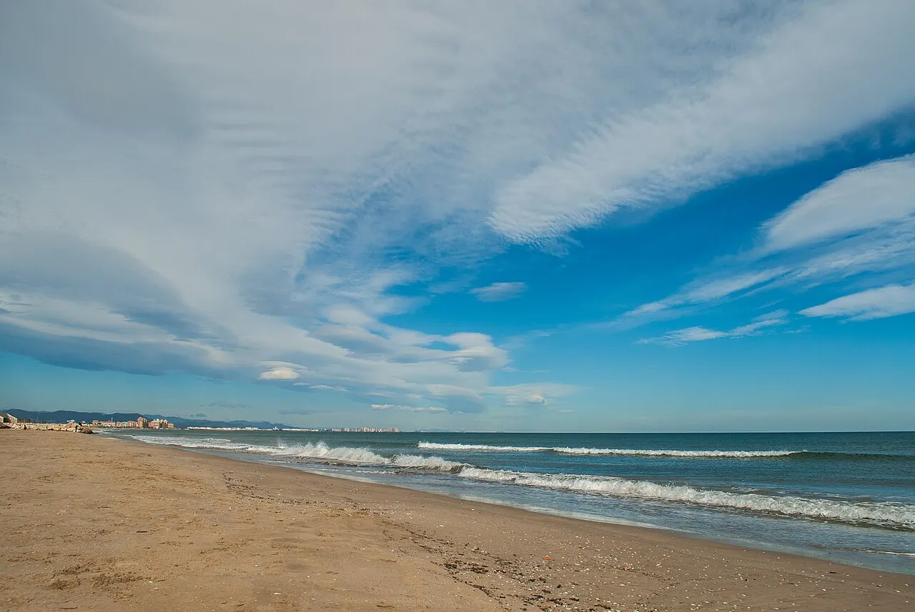 A Playa de la Malvarrosa, uma faixa larga de areia dourada com ondas pequenas, sob céu azul riscado de nuvens.