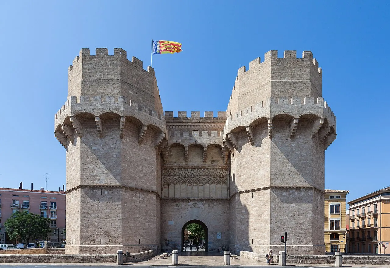 As Torres de Serranos, porta medieval de pedra com duas torres poligonais ameadas e uma bandeira no alto, sob céu azul.