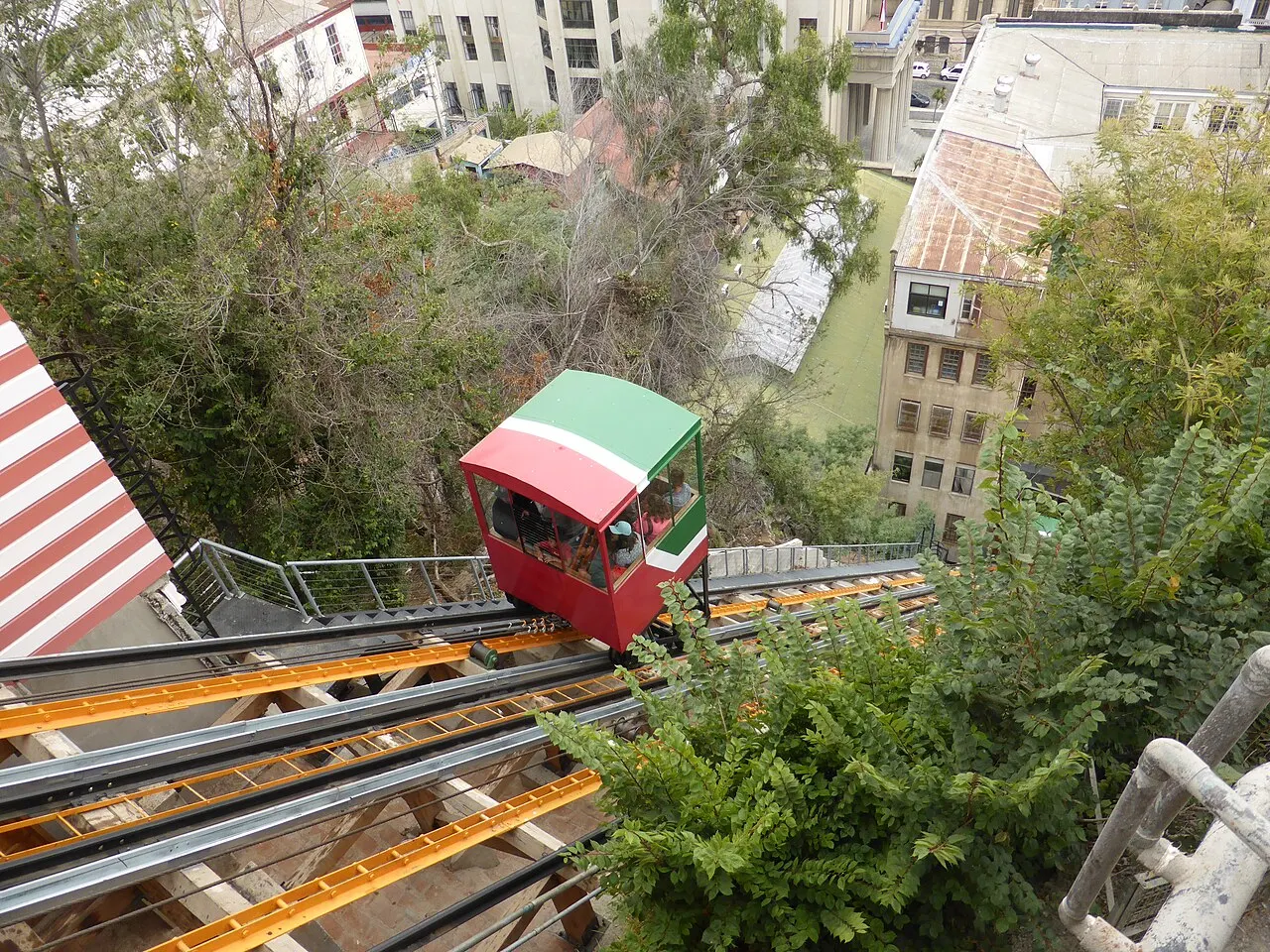 Cabine vermelha e verde do funicular Ascensor El Peral descendo trilhos amarelos entre árvores, com telhados e prédios de Valparaíso abaixo.