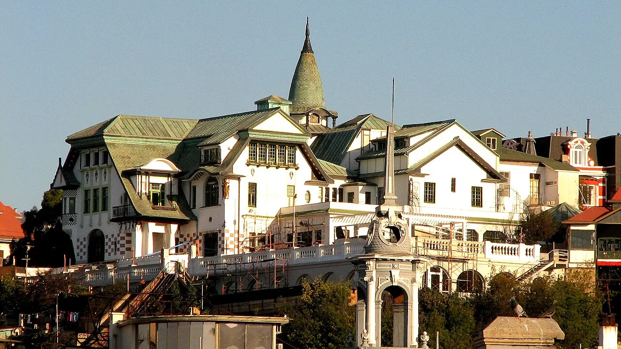 Mansão art nouveau do Palacio Baburizza, com telhados verdes inclinados, uma torre cônica e varandas brancas, iluminada por sol lateral.