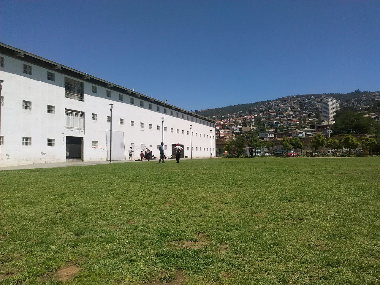 Bloco comprido e branco da antiga prisão de Valparaíso, com janelas gradeadas em duas fileiras, sobre uma esplanada gramada, e as casas coloridas dos cerros subindo a encosta ao fundo.