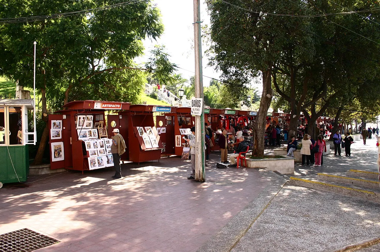 O Paseo 21 de Mayo, com quiosques vermelhos de venda, pessoas caminhando e árvores dando sombra.