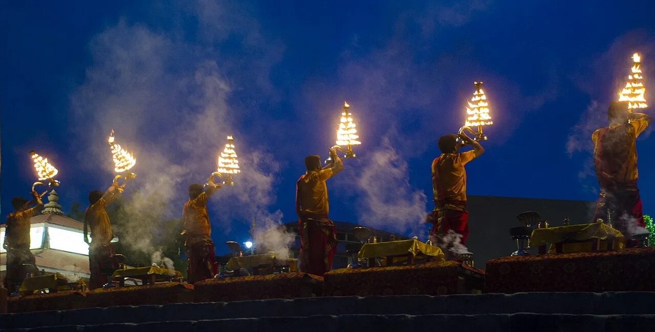 Sacerdotes de roupa cor de açafrão erguem candelabros acesos na plataforma do ghat, com fumaça de incenso contra o céu azul do amanhecer.