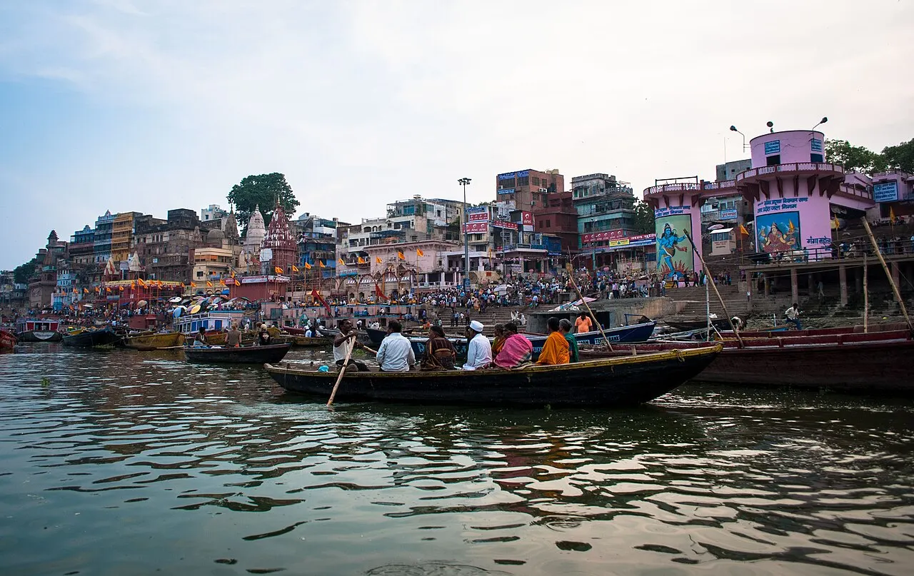Barcos de madeira lotados de passageiros no rio Ganges em frente ao Dashashwamedh Ghat, com multidão nas escadarias e prédios coloridos ao fundo.