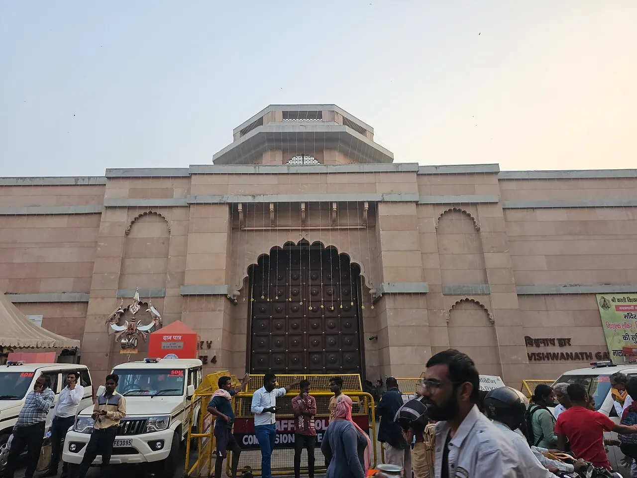 Fachada de pedra rosada do templo de Kashi Vishwanath, com um portal em arco ornamentado e um andar octogonal no alto, carros e gente na rua em frente.