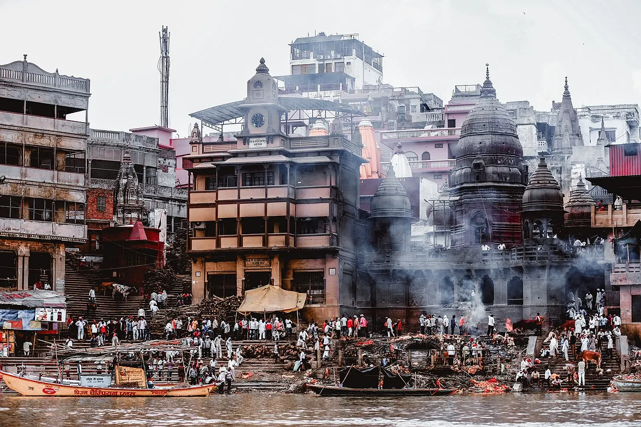 Construções escurecidas por fumaça no Manikarnika Ghat, à beira do rio Ganges, com fumaça subindo entre os prédios e pessoas reunidas nas escadarias.