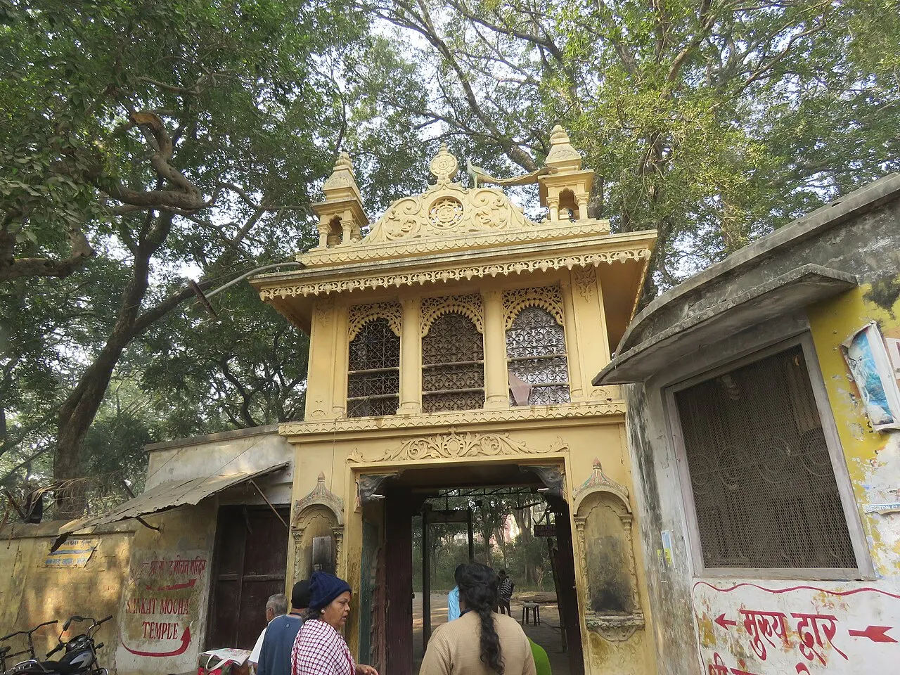 Portal amarelo de entrada do Templo Sankat Mochan, entre árvores, com visitantes passando pela porta.