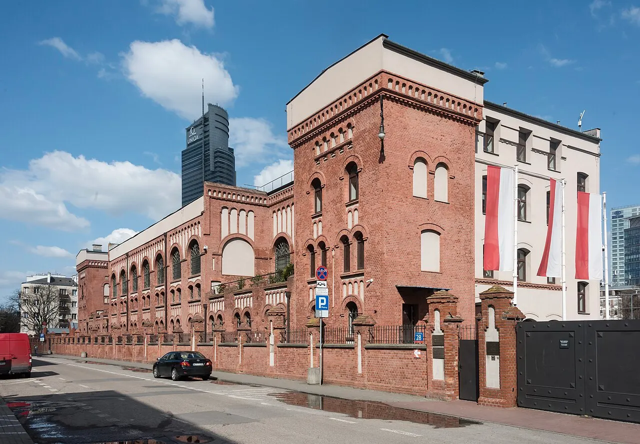 Fachada de tijolo vermelho do Muzeum Powstania Warszawskiego, com janelas em arco, bandeiras da Polônia penduradas e uma torre de vidro moderna ao fundo.