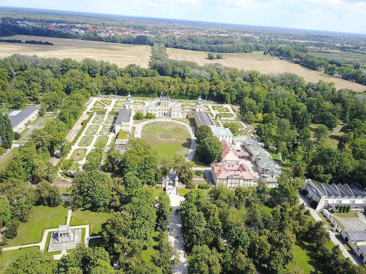 Vista aérea do palácio barroco de Wilanów, claro e de duas alas, com os canteiros geométricos dos jardins de um lado, o gramado oval na frente e o bosque em volta.