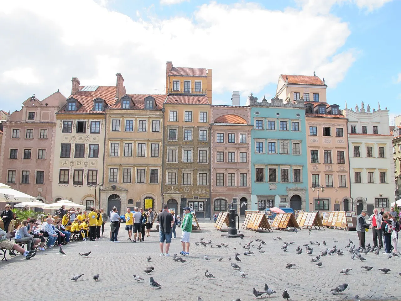 Fileira de casas coloridas de fachada estreita na Rynek Starego Miasta, com pombos no chão de pedra e gente sentada em bancos à sombra.