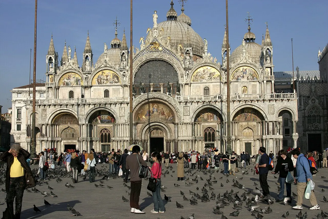 Fachada ornamentada da Basílica de San Marco, com cúpulas bizantinas, mosaicos dourados sobre os portais e uma multidão de pessoas e pombos na praça.