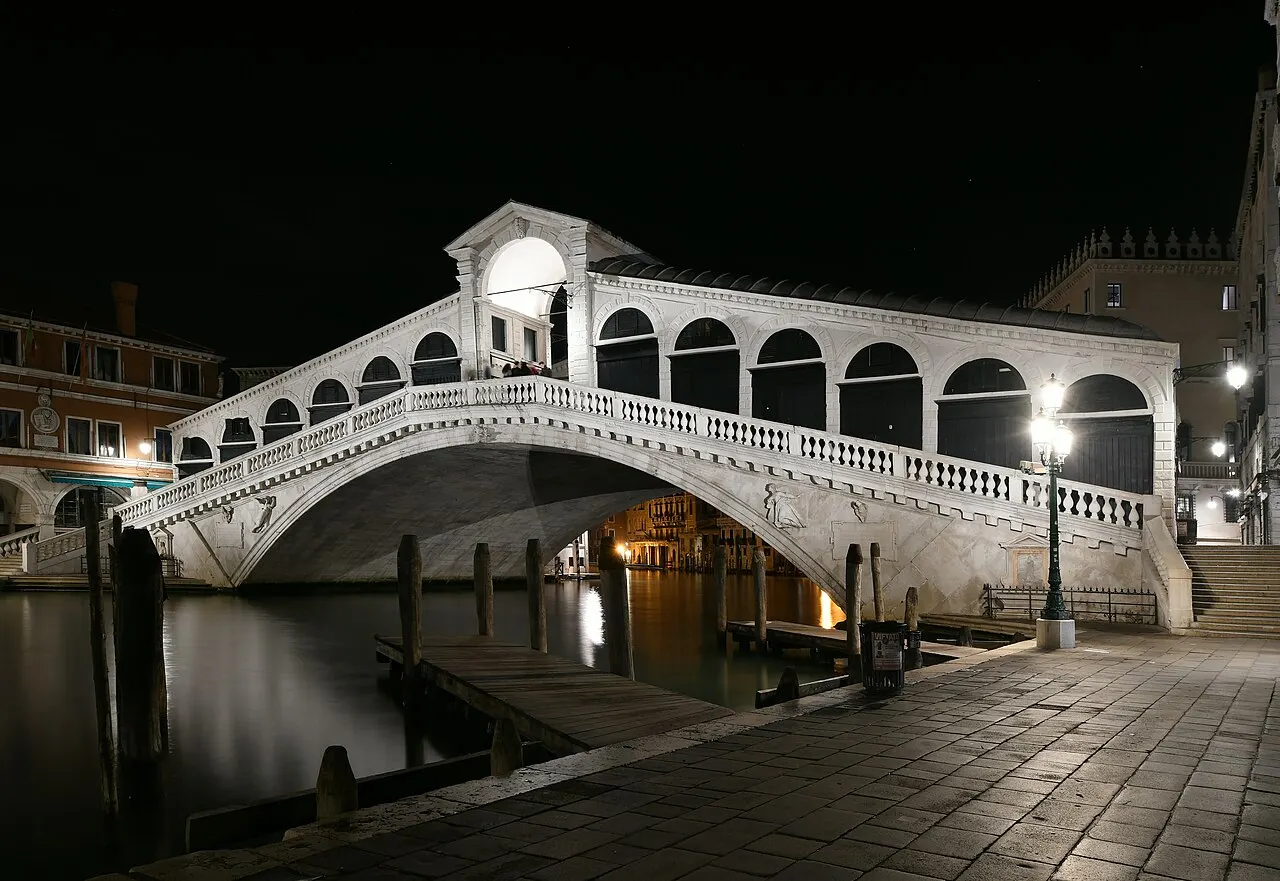 Ponte de Rialto, em Veneza, com o arco de pedra e as lojas em cima, iluminada à noite sobre o Grande Canal.