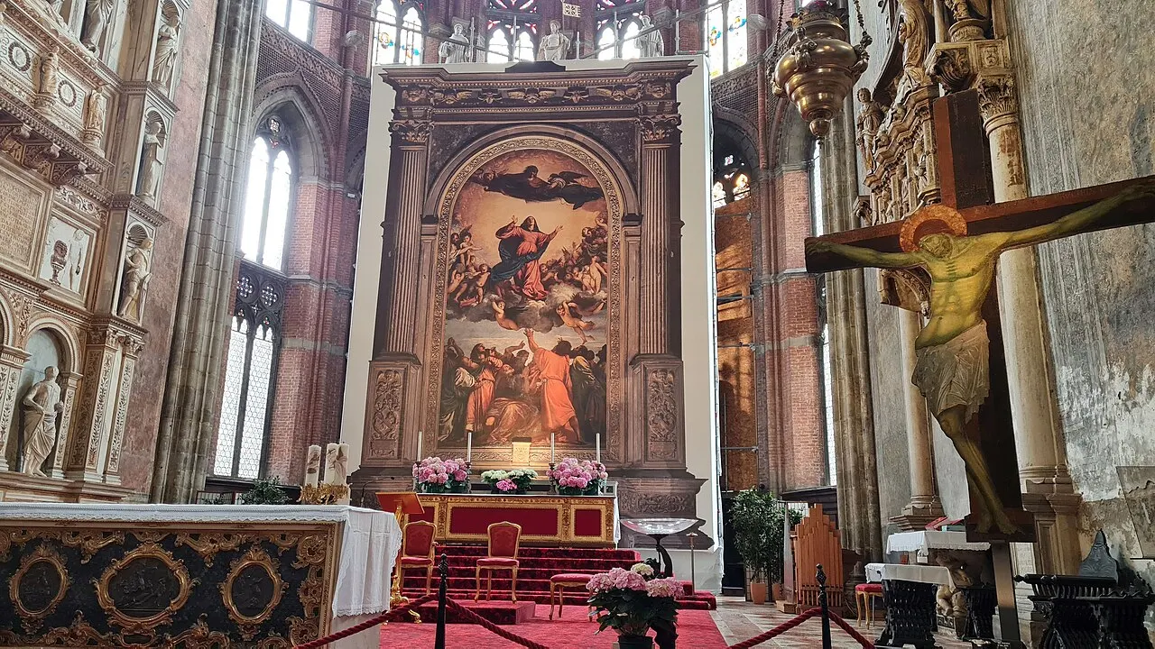 Interior gótico de tijolo da basílica dos Frari com a Assunção de Ticiano sobre o altar-mor, flores no altar, degraus vermelhos e um crucifixo de madeira suspenso à direita.