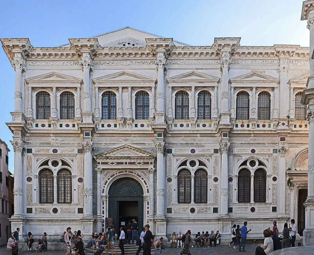 Fachada de mármore branco da Scuola Grande di San Rocco, com colunas, janelas ornamentadas e pessoas na entrada.