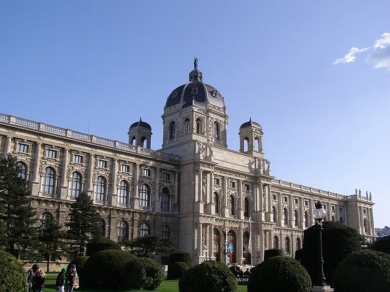 Fachada monumental do Kunsthistorisches Museum, com uma cúpula central coroada por uma estátua, torres menores nas laterais e jardim aparado em frente.