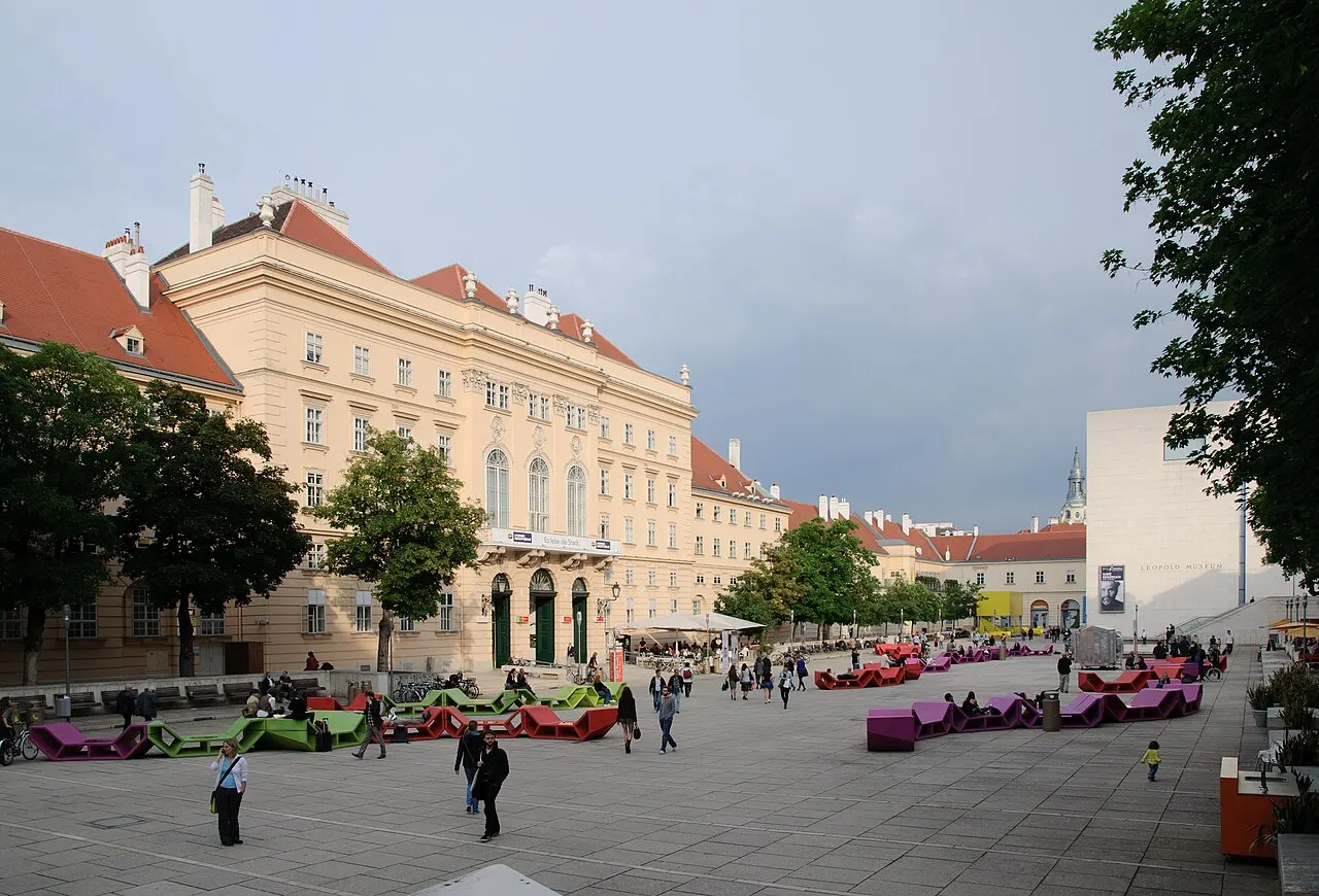The courtyard of the MuseumsQuartier, with people and brightly colored furniture, surrounded by Baroque buildings.
