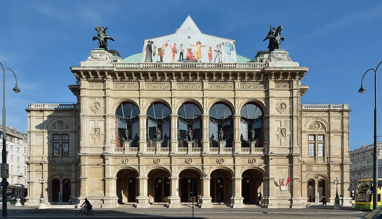 Fachada da Ópera Estatal de Viena, com arcadas, janelas em arco e estátuas no alto, sob céu azul.