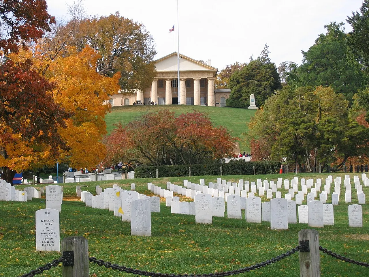 Lápides brancas em fileiras no Cemitério Nacional de Arlington, com árvores de outono e a Arlington House no alto.
