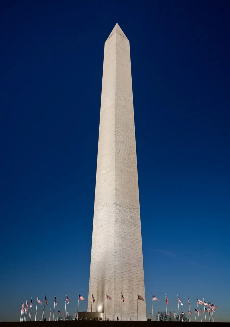 Obelisco do Washington Monument ao entardecer, contra o céu azul-escuro, com bandeiras na base.