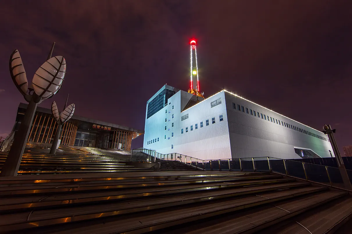 Prédio industrial branco iluminado à noite, com chaminé vermelha e verde ao fundo.
