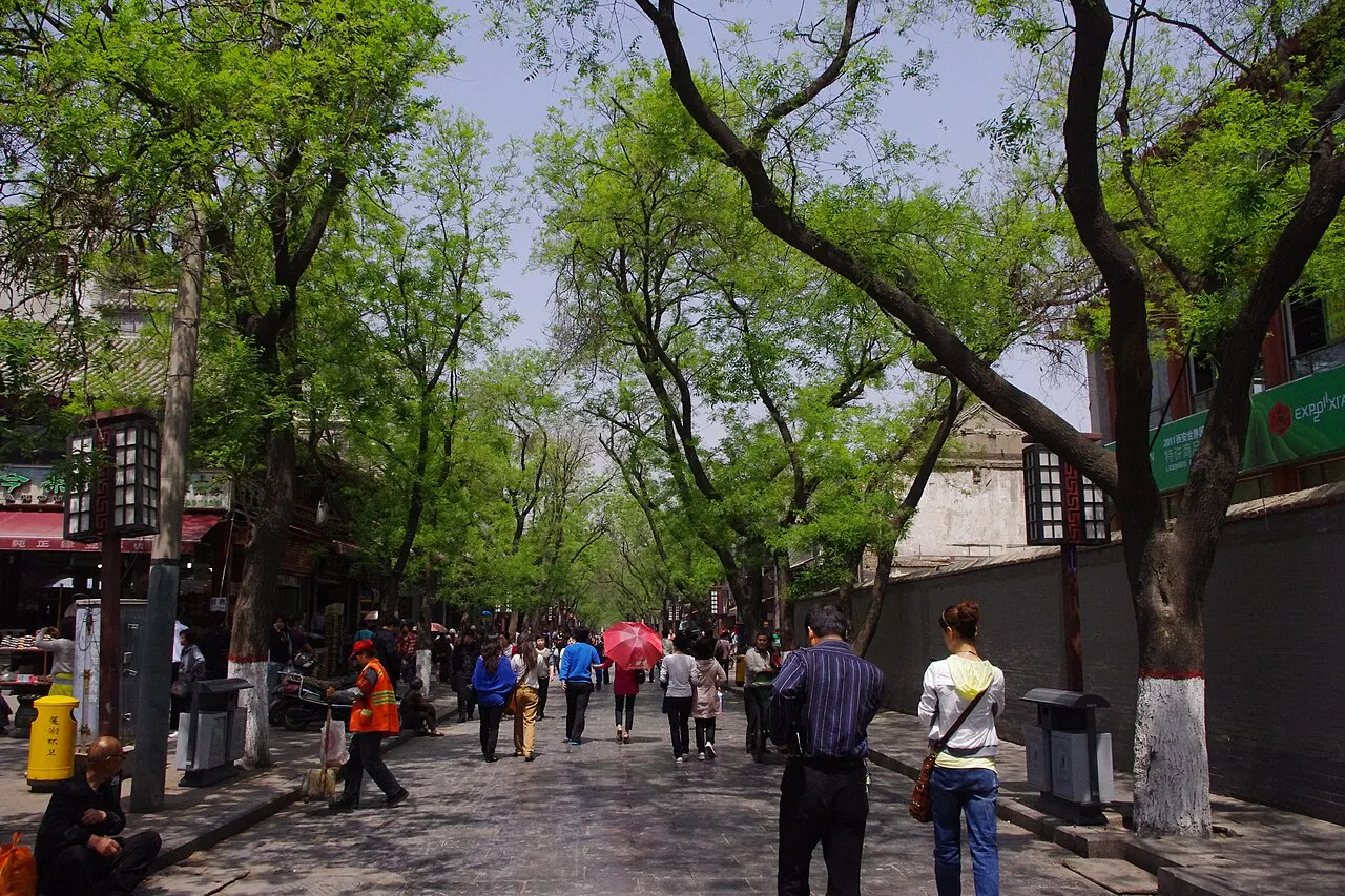 Beiyuanmen street in Xi'an's Muslim Quarter, paved in stone under a line of tall trees, with shops on the left, a grey wall on the right and people walking.